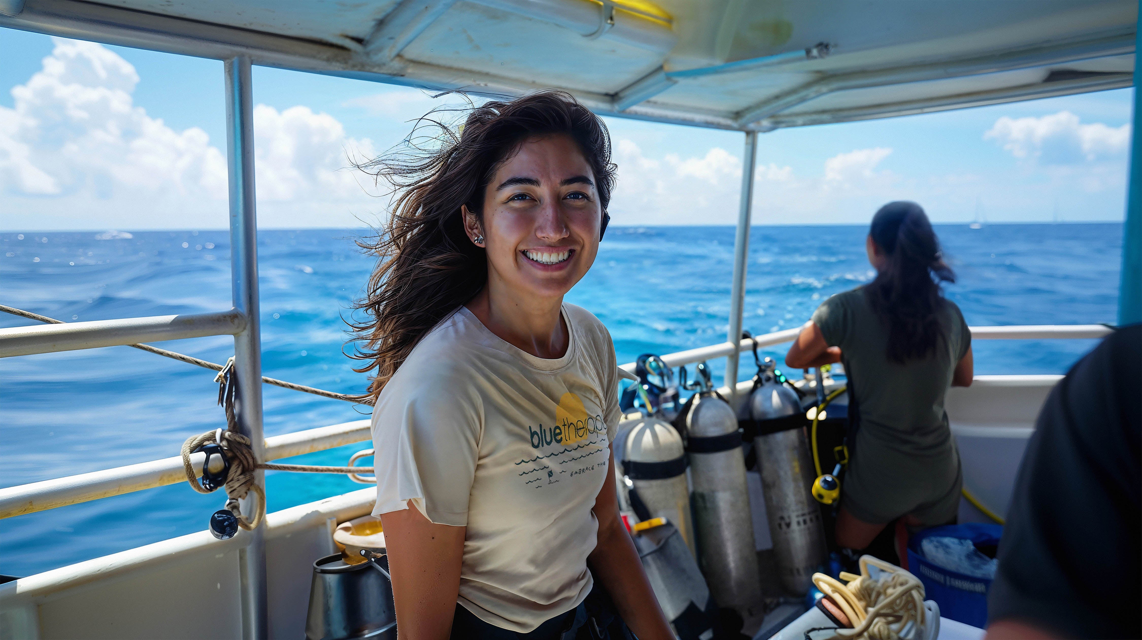 Abbie on dive boat wearing BlueTherapy Embrace the Blue t-shirt, wind in hair, smiling with ocean horizon, dive gear and tanks in background