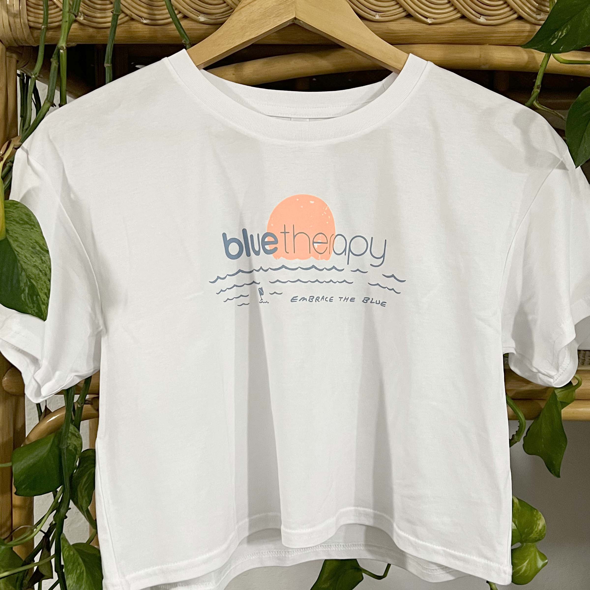 White BlueTherapy 'Embrace the Blue' crop tee with blue and orange text design, displayed on a hanger with foliage in the background.