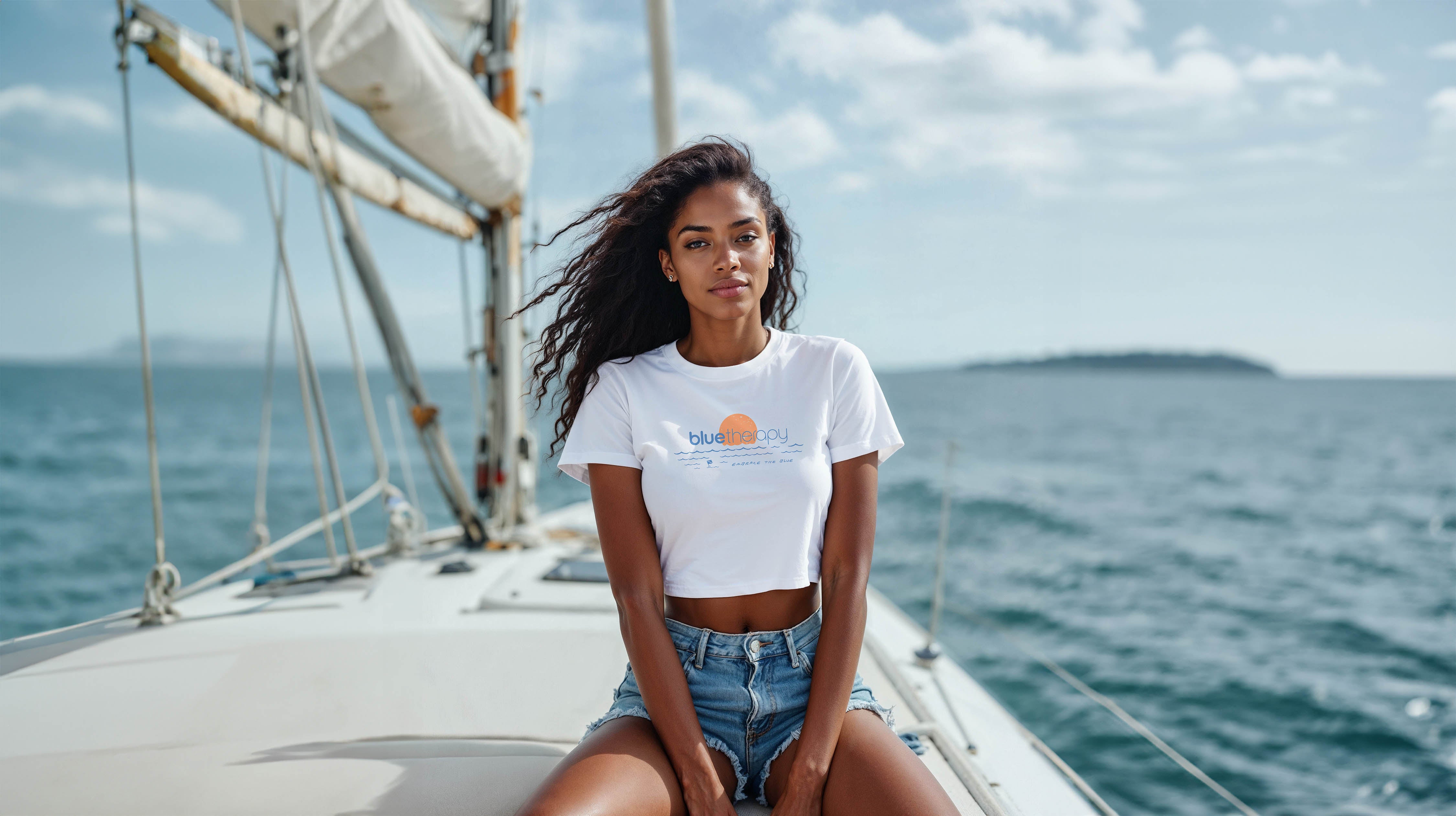 Model relaxing on sailboat in BlueTherapy Embrace the Blue crop tee featuring blue waves and sun design, scenic ocean and sky background