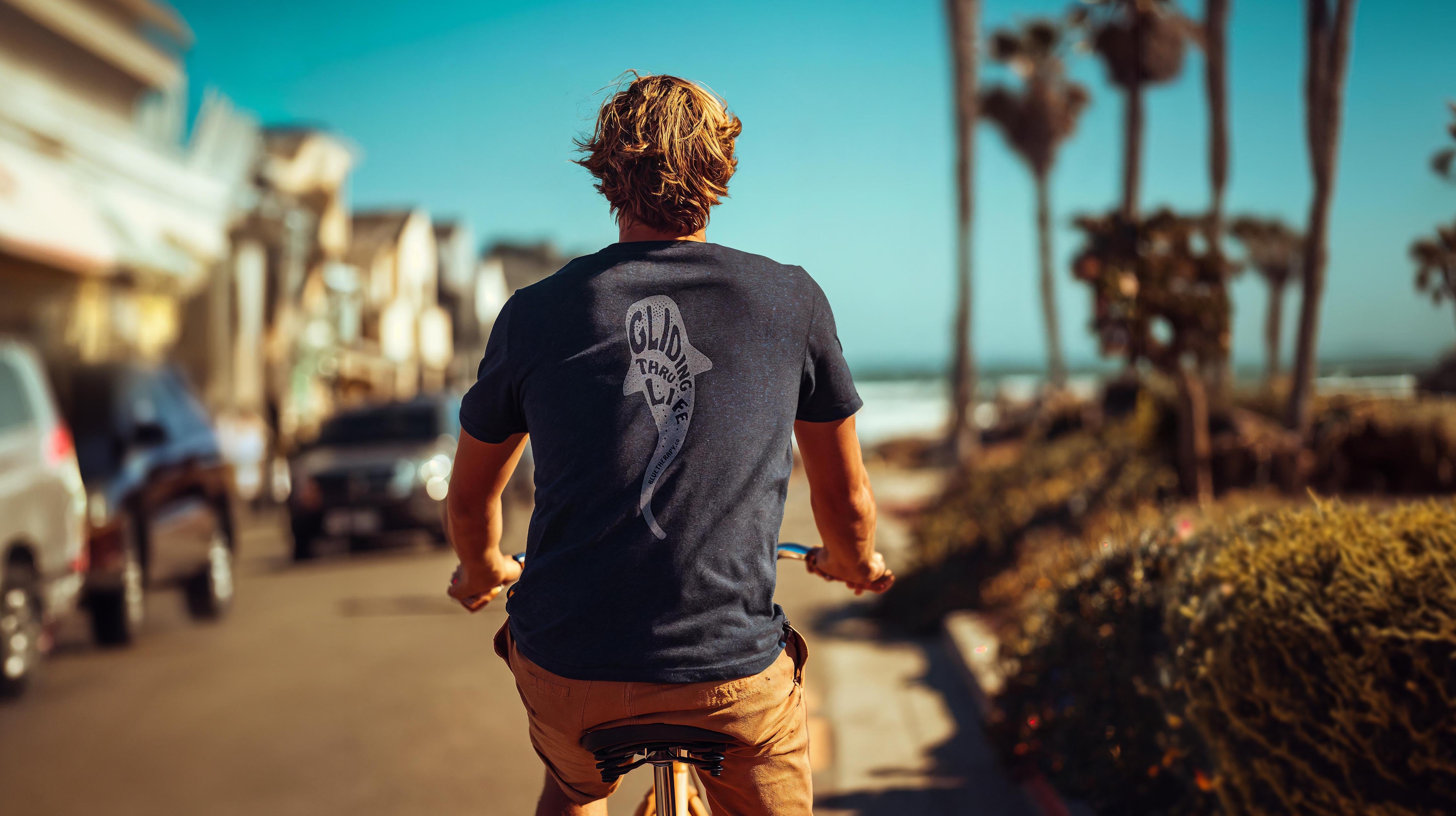 Man riding beach cruiser in BlueTherapy Gliding Whale Shark tee riding along sunny beachfront path, full back view showing detailed 'Gliding Thru Life' whale shark illustration and ocean view