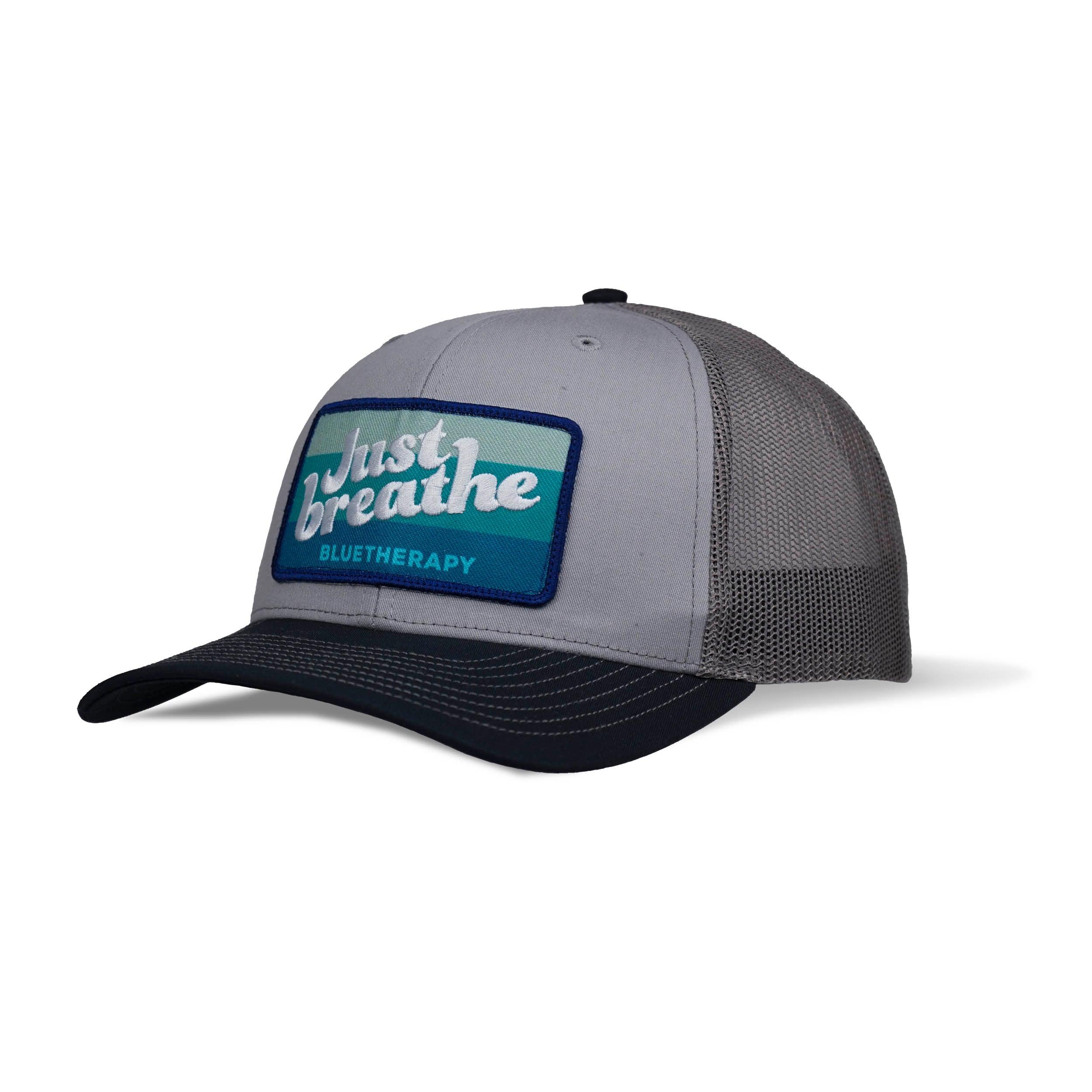 3-qtr view of trucker hat with a navy curved brim and a white embroidered text 'Just Breathe' on a blue blended patch, with a charcoal mesh back and an adjustable snap.
