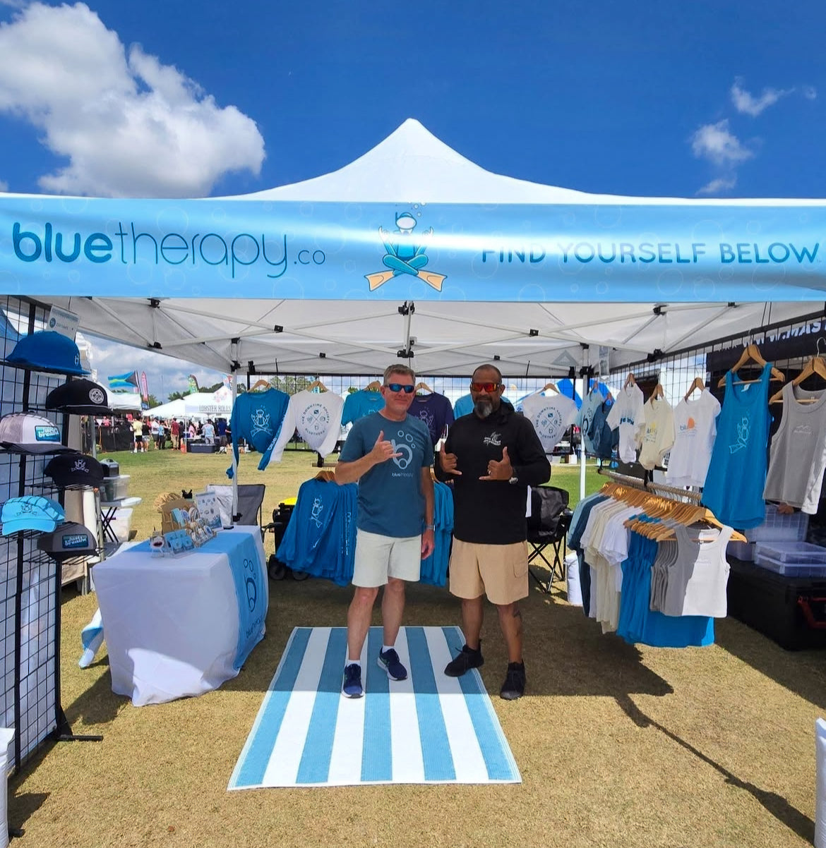 Tom from BlueTherapy and Rafael with Military Recreational Divers standing under a blue 'bluetherapy.co' tent at an outdoor event.
