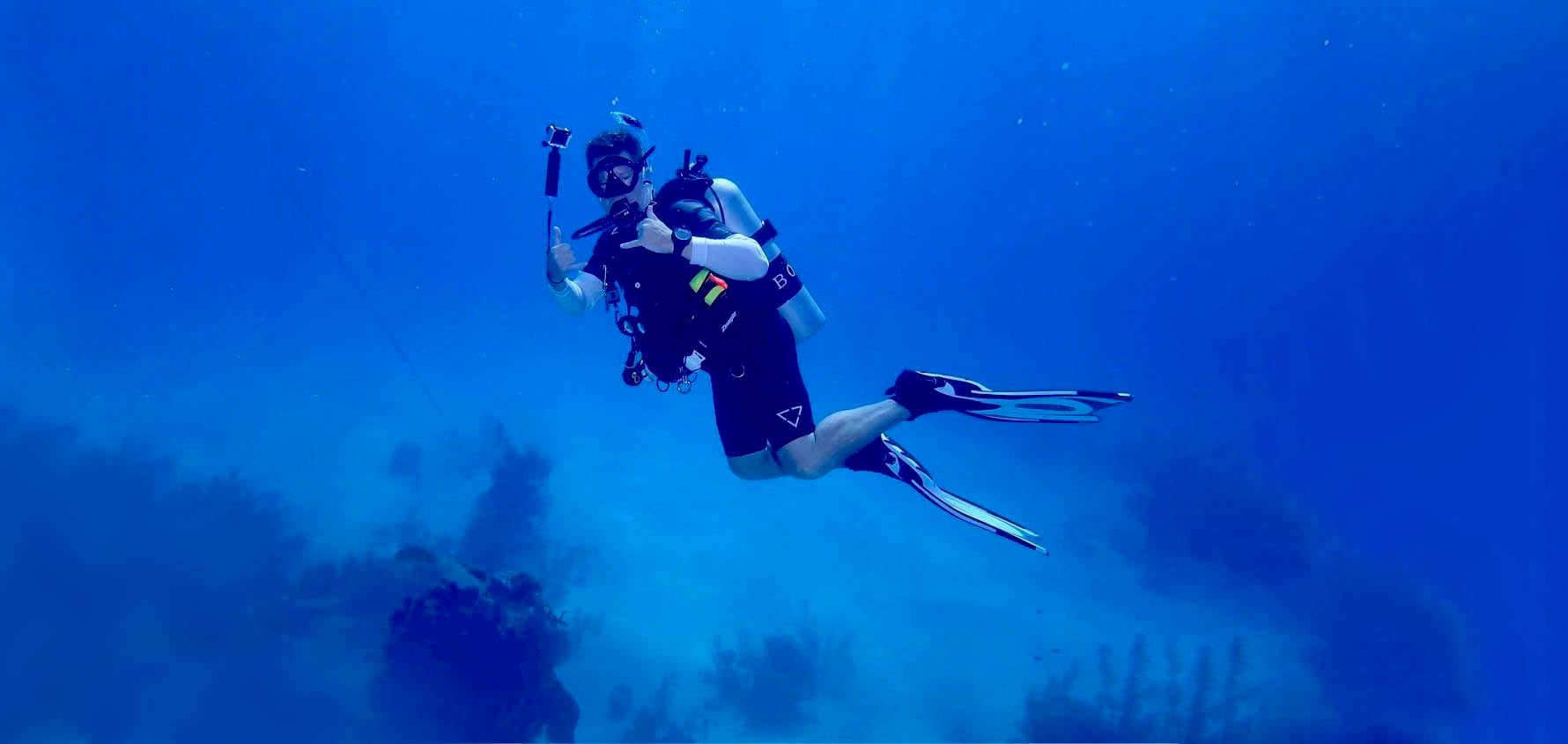 BlueTherapy founder Tom Sloan underwater during a SCUBA dive, giving a shaka sign while floating weightlessly in clear blue ocean with coral reef below, capturing the 'Just Breathe' zen therapy moment
