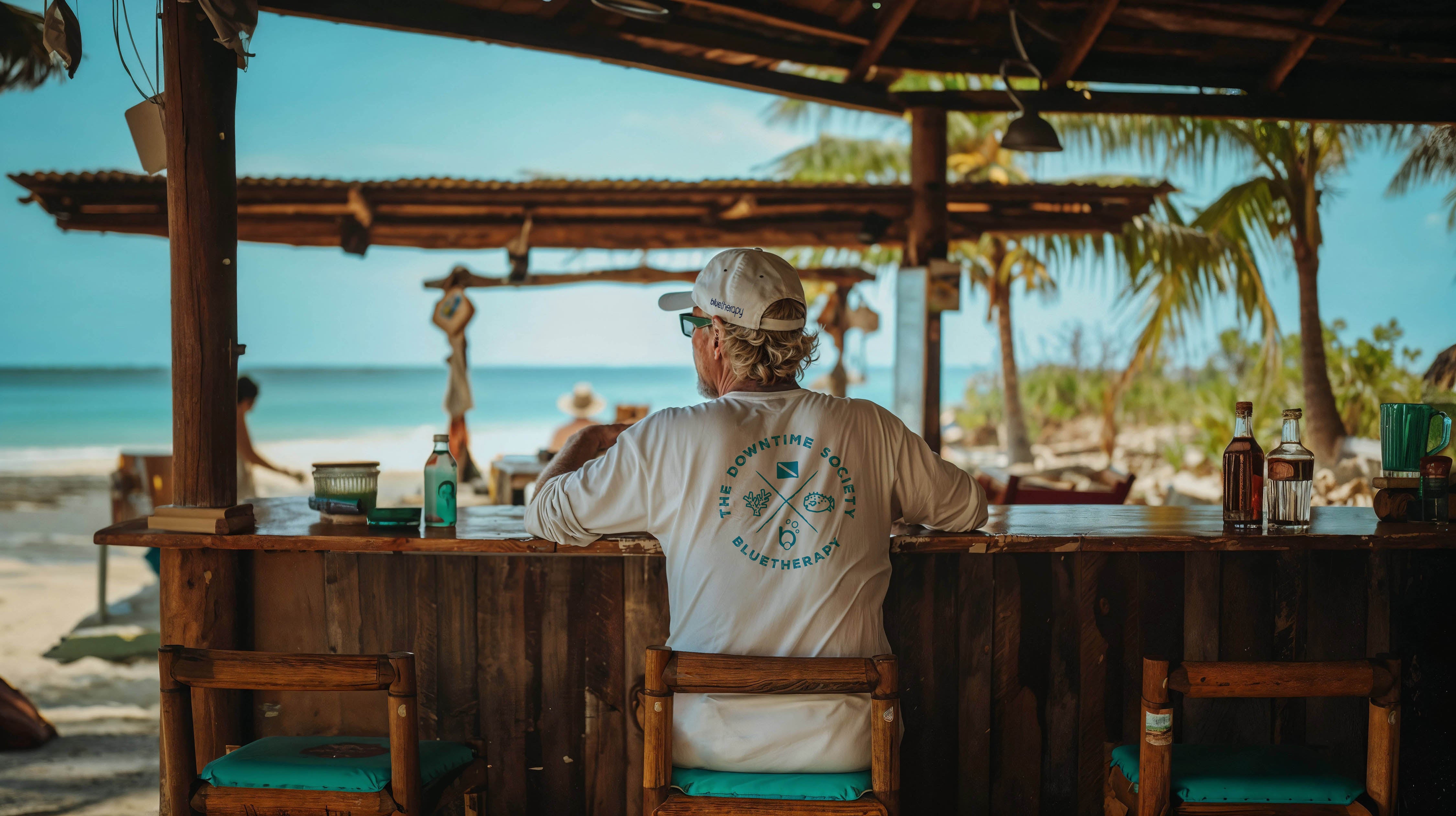 Al in BlueTherapy Downtime Society UPF longsleeve sunshirt sitting at tropical beach bar counter, back view overlooking ocean, palm trees, and horizon