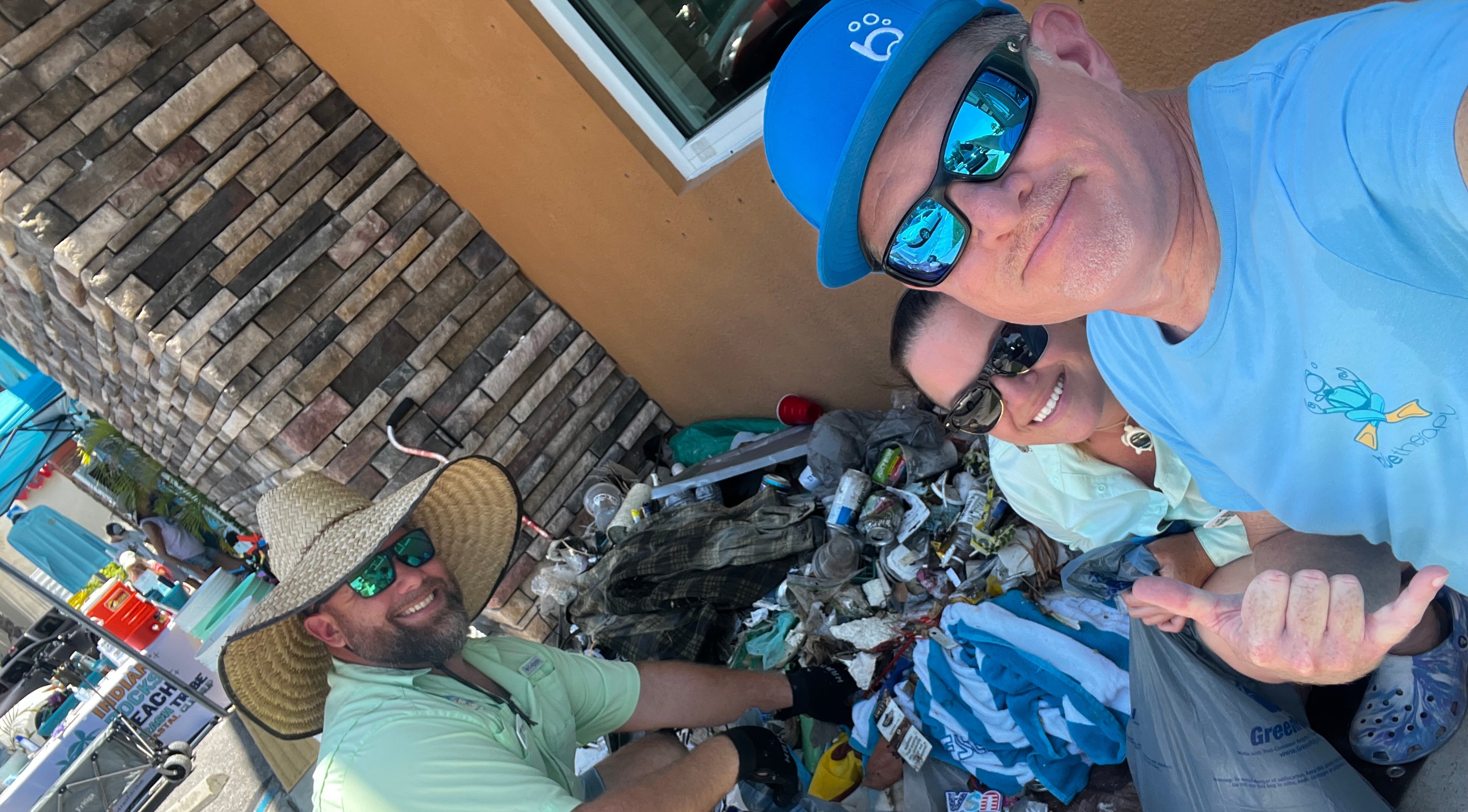 three people outdoors at the indian rocks beach clean up, Tom from BlueTherapy wearing a blue bubble b hat and sunglasses, and Jenn and Don from IRB Trash Tribe, with a pile of trash picked up from the area including bottles and bags.