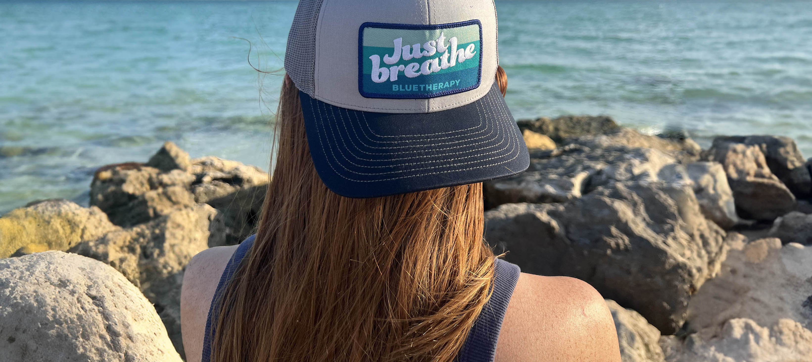 Madi looking out at turquoise ocean wearing BlueTherapy Just Breathe trucker hat, back/side view with 'Just Breathe' patch on front, rocky beach foreground