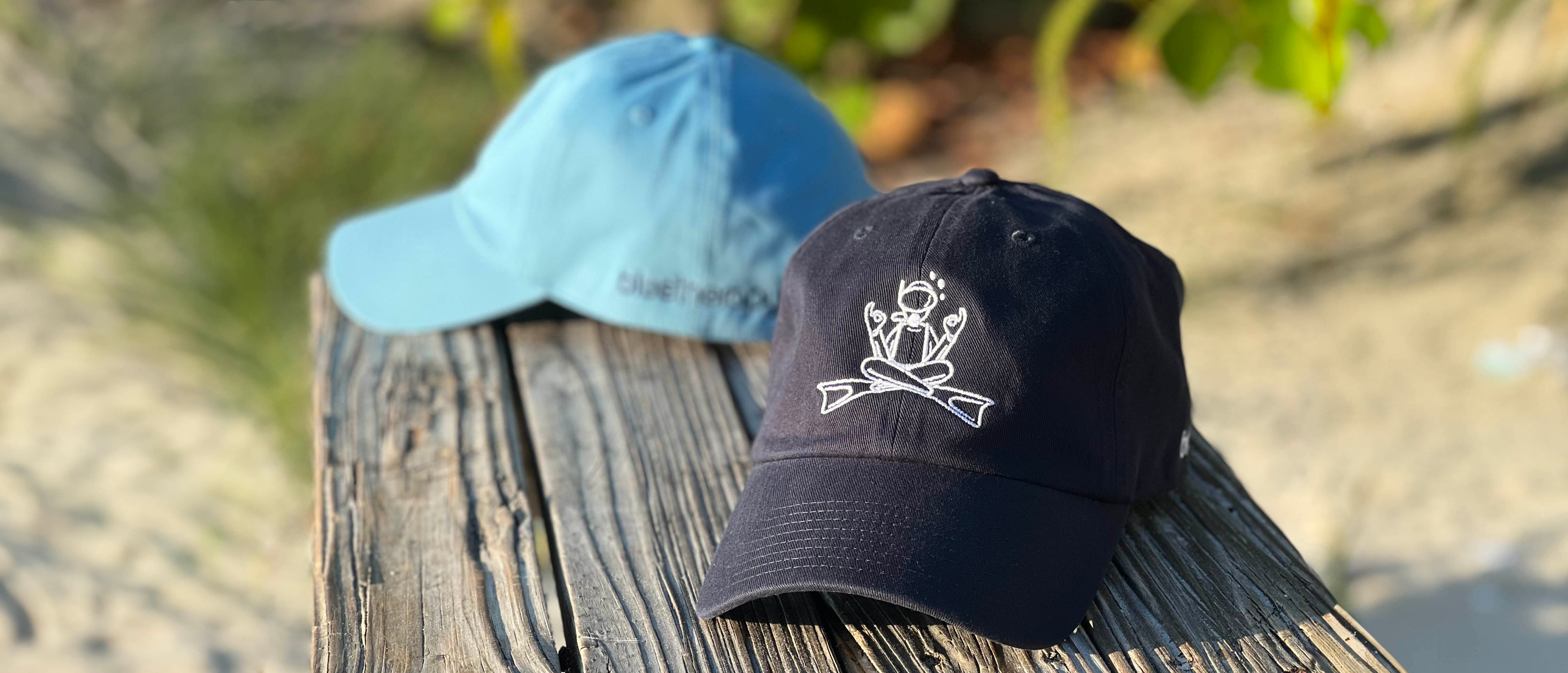 BlueTherapy Zen dad hats displayed on wooden bench outdoors, light blue and navy caps with minimalist diver in lotus pose embroidery, tropical plant background